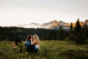 couple sits and kisses in field looking over Mount rainier landscape