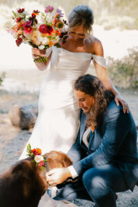 bride holds bouquet while groom puts flower collar on dog