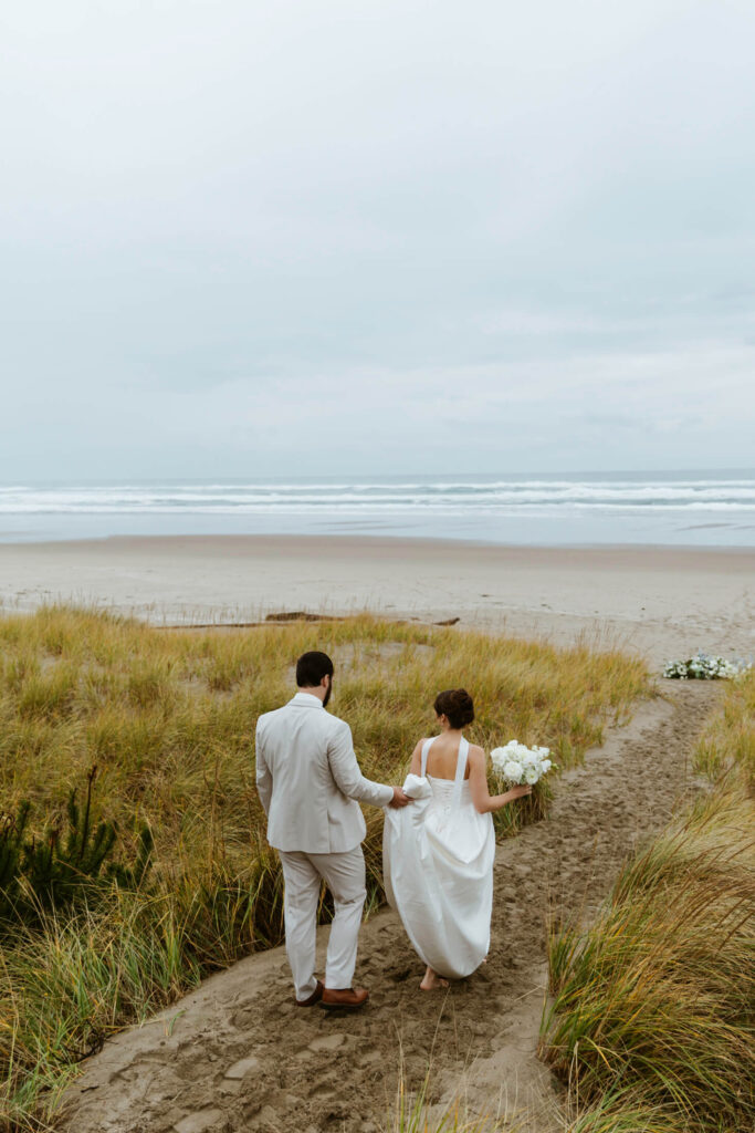 Groom holds his brides train as she walks down a sand aisle for their vow renewal
